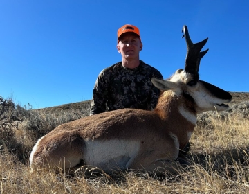 A hunter in a camouflage shirt and orange cap kneels beside a large pronghorn antelope, basking in the Wyoming sun after a successful SNS-guided hunt.